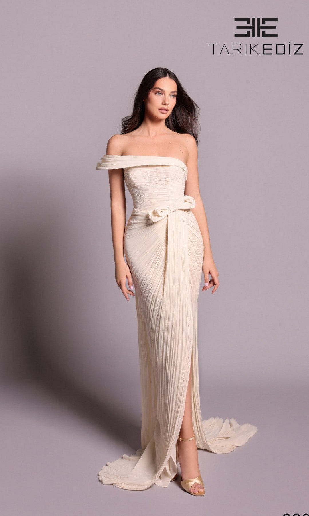 A woman models the Tarik Ediz 99095 long formal dress in vanilla, featuring an off-shoulder neckline, pleats, and a waist bow, standing against a plain background with the Tarik Ediz logo visible.